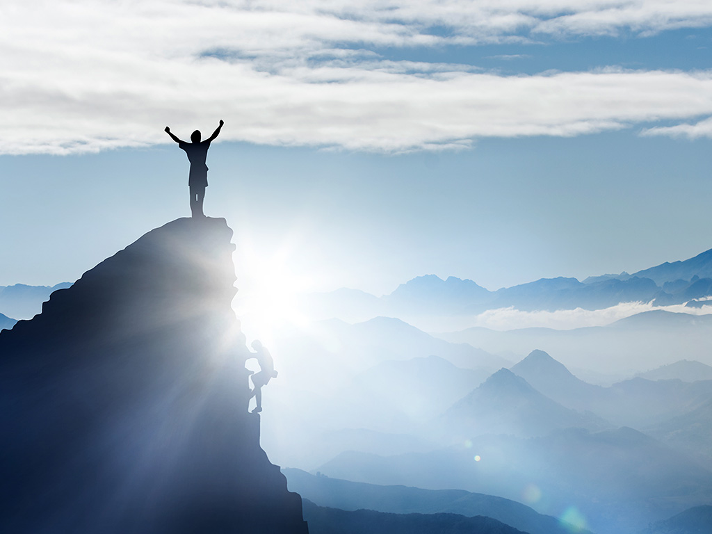 Eine Person steht auf einem Gipfel eines Berges, hebt die Arme in die Luft und blickt in die Sonne.